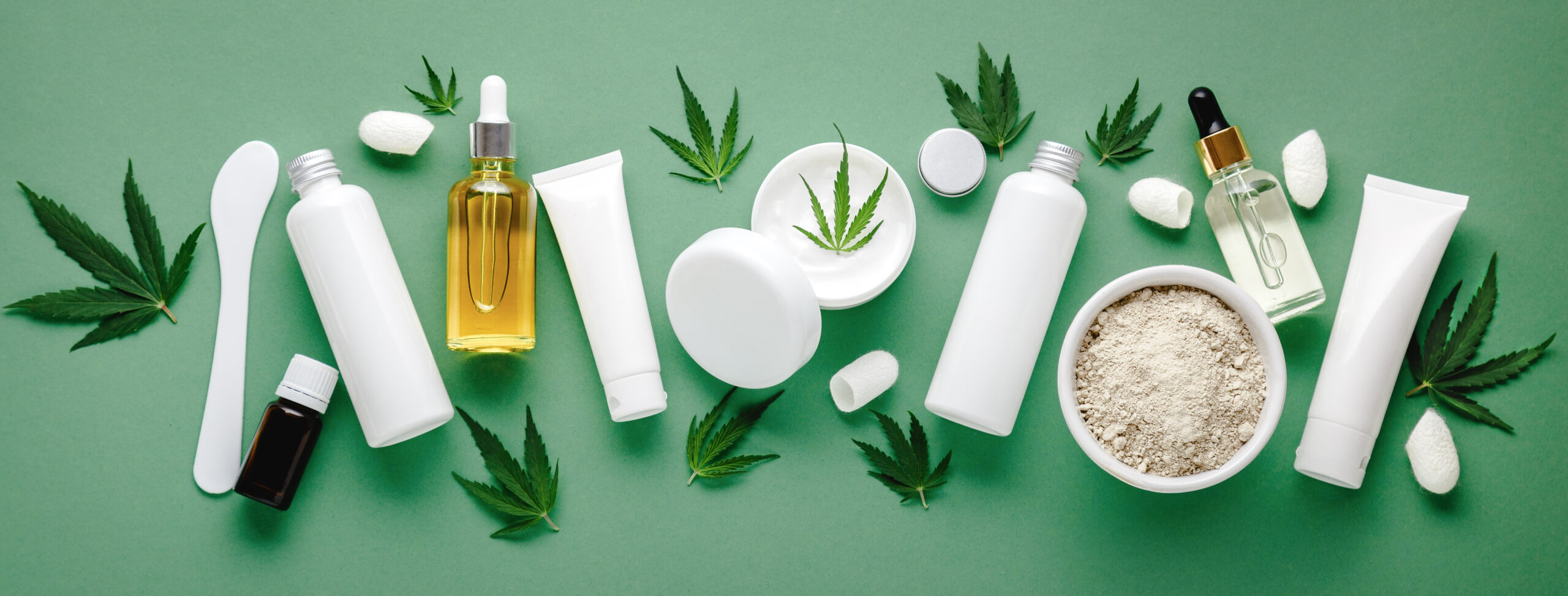 Set of hemp skin care cosmetics in white mockup packaging. Moisturizing cream, Serum, lotion, CBD oil, essential oil cannabis leaves. Flat lay long web banner copy space. on green background.